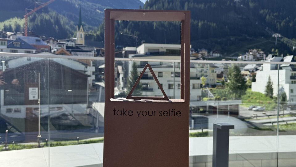 A self-portrait frame with the prompt "take your selfie" stands in front of a mountain landscape. In the background, buildings and green areas are visible.