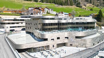 A modern hotel building in a mountainous area. Surrounded by green meadows and traditional chalets.