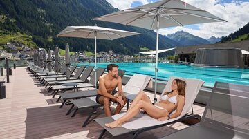 A relaxed day by the pool with loungers and umbrellas. A couple enjoys the sun and reads a book.