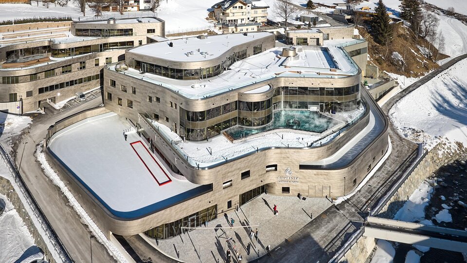 A modern building in a snow-covered landscape with an ice rink accessible all year round. The architecture is elegant, surrounded by picturesque mountains and vacation homes.
