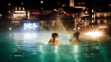 Two women are enjoying drinks in the illuminated swimming pool at night. The atmosphere is relaxed, and the city lights are shining in the background.