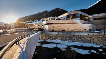 A modern building in an alpine setting. The sun shines over the mountains and reflects on the facade.