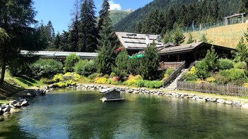 An idyllic pond surrounded by green trees and a mountainous landscape. In the background, a wooden house is visible, harmoniously integrated into nature.