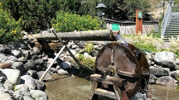 A waterwheel powered by flowing water stands beside a river. In the background, green plants and a wooden house can be seen.