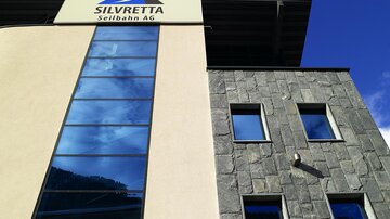 A modern building with a glass facade and a large sign of the company Silvretta Seilbahn AG. The sky is clear and blue, creating a pleasant atmosphere.