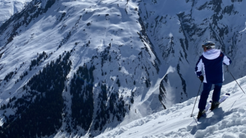 A skier stands on a snowy slope with high mountains in the background. The sky is clear and blue.