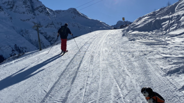 A skier is going down a snow-covered slope while a dog sits next to him. The sun is shining clearly in the blue sky.