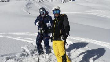 Two skiers are standing in fresh snow under a clear sky. Snow-covered mountains can be seen in the background.