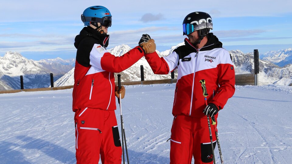 Two skiers in red suits are standing on a snowy summit. In the background, the majestic mountains are visible.
