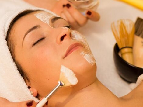 Eine Frau erhält eine Gesichtsbehandlung mit einer Maske auf ihrem Gesicht. Um sie herum sind verschiedene Beauty-Werkzeuge und eine Schüssel sichtbar.