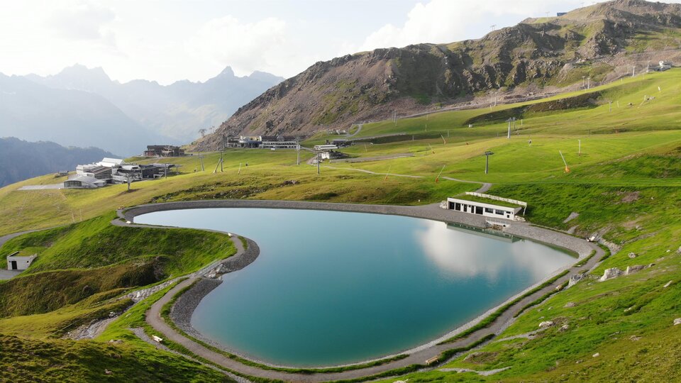 A picturesque mountain lake surrounded by green meadows and high mountains. The water's surface reflects the landscape.