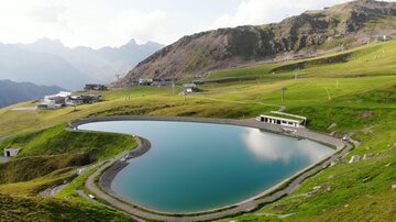 A quiet heart-shaped mountain lake, surrounded by green meadows and mountains. In the background, modern buildings are visible.