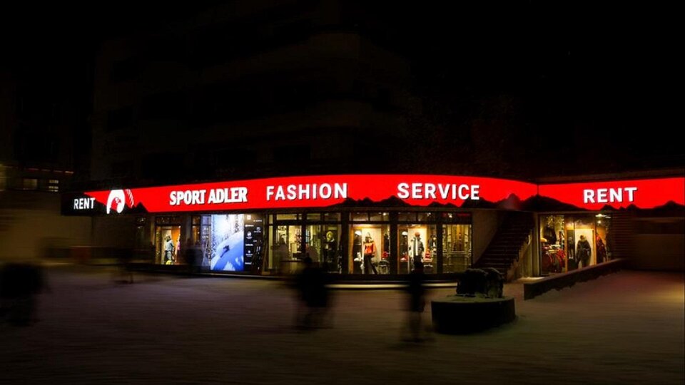 Ein Geschäft mit leuchtendem, rotem Schild, das "Sport Adler" und "Fashion Service" anzeigt. Die Szene ist nachts, und es sind verschwommene Gestalten im Vordergrund zu sehen.