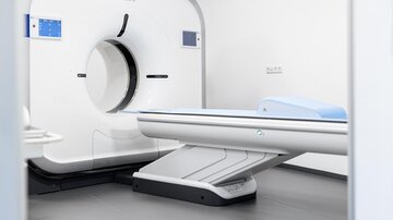 A modern CT scanner stands in a bright, minimalist room. The equipment is designed in white and gray, appearing clean and technologically advanced.