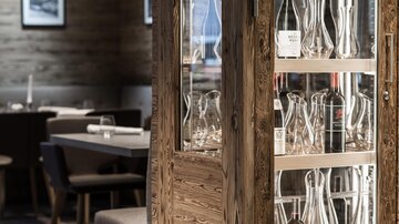Ein eleganter Restaurantbereich mit einem Glasregal aus Holz. Die Einrichtung ist modern und stilvoll, mit gemütlichen Tischen.