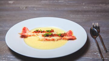 Ein elegantes Gericht auf einem weißen Teller mit zwei gefüllten Teigtaschen und einer cremigen Sauce. Das Gericht ist mit frischen Kräutern und einer roten Sauce garniert.