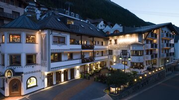 Ein schönes Hotelgebäude im alpinen Stil, umgeben von Bergen. Die Fassade ist in sanften Farben gestaltet und abends sanft beleuchtet.