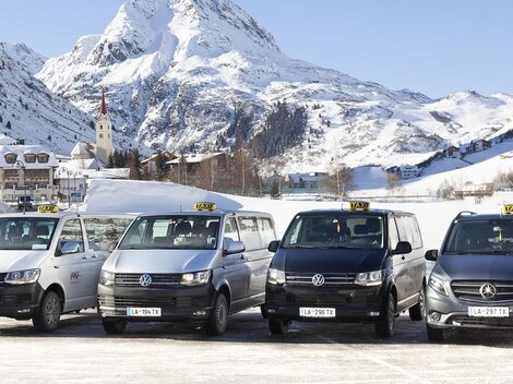A group of taxis stands in front of a snow-covered mountain landscape. In the background, majestic mountains and a small town can be seen.