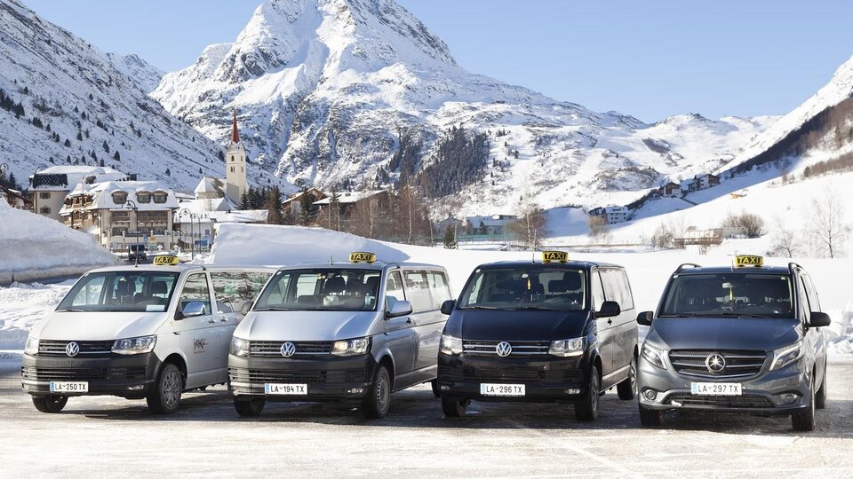 A group of taxis stands in front of a snow-covered mountain landscape. In the background, majestic mountains and a small town can be seen.