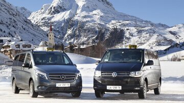 Two taxis stand in the snow in front of an impressive mountain landscape. In the background, snow-covered mountains and a small village can be seen.