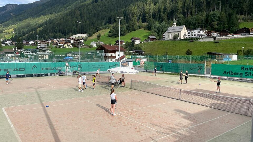 A tennis court with several players and a mountainous landscape in the background. The surroundings are green with typical Alpine architecture.
