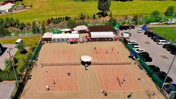 A tennis facility with multiple courts and players. In the background, there are buildings and vehicles, surrounded by green landscape.