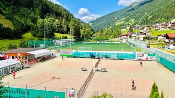 A beautiful tennis court in a green landscape with mountains in the background. Some players are active while others are sitting on the bench.