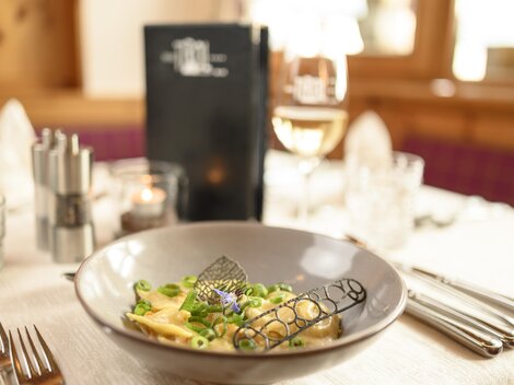 A stylishly set table with a plate of pasta and fresh vegetables. In the background, there is a drink and a menu card visible.