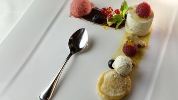 An artfully arranged dessert with various scoops of ice cream, fresh berries, and a creamy ingredient. A silver spoon complements the elegant presentation on the white plate.