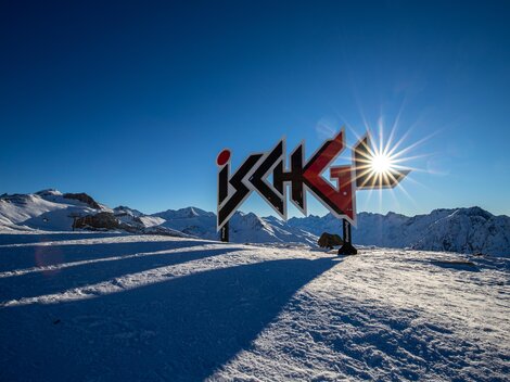 A beautiful viewpoint in Ischgl with a large sign and snow-covered mountains. The sun shines in the clear blue sky.