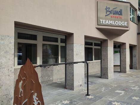A modern building with a sign that says "Teamlodge." In front of the entrance is another sign featuring sporty motifs.