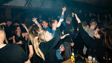 Eine lebendige Partyszene mit tanzenden Menschen in einem dunklen Raum. Die Atmosphäre ist energiegeladen, und viele haben Getränke in der Hand.