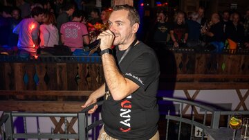 Ein Mann mit Mikrofon steht in einem Club und singt. Die Menge ist im Hintergrund sichtbar und hat Spaß.