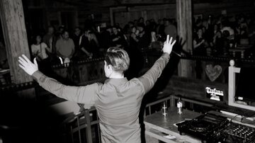 Ein DJ steht hinter dem Pult und hebt die Arme, während das Publikum ihn anfeuert. Die Szene ist dunkler, mit stimmungsvollem Licht und einer lebhaften Atmosphäre.