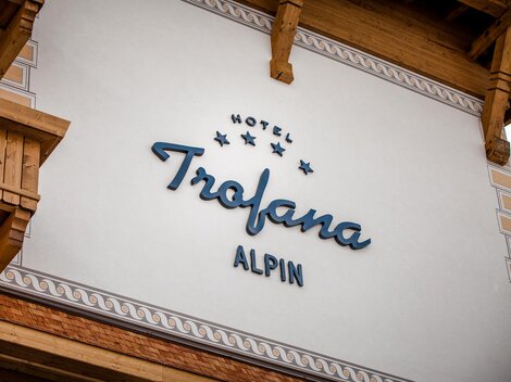 A hotel sign with the name "Trofana Alpin" is mounted on the wall. The façade features rustic wooden elements and an elegant font.