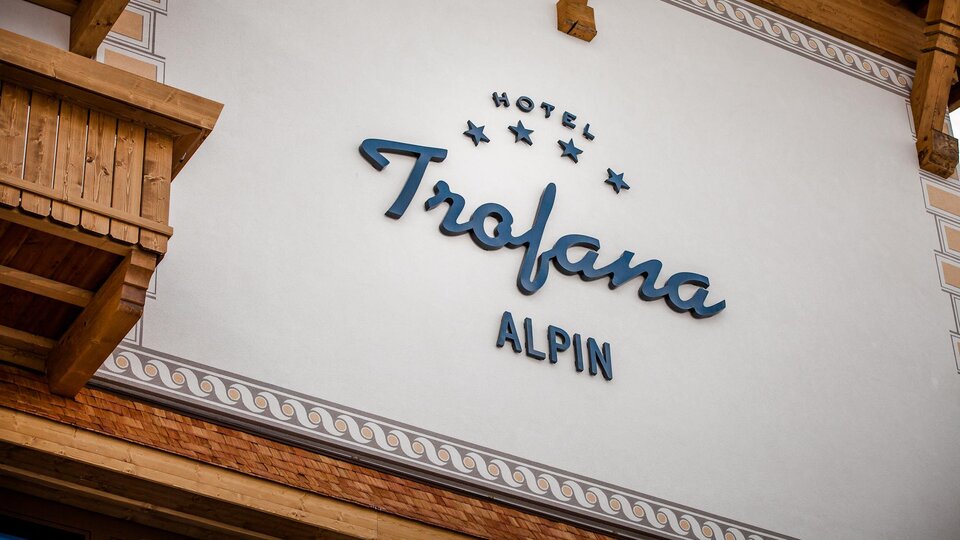 A hotel sign with the name "Trofana Alpin" is mounted on the wall. The façade features rustic wooden elements and an elegant font.