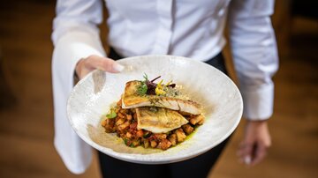 A well-arranged plate with fried fish and colorful vegetables. The service is presented by a person in a white shirt.
