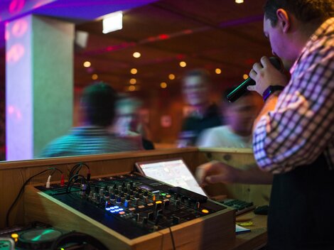 A DJ stands behind a mixing console and holds a microphone in hand. The colorful lights and the dancing crowd create a vibrant party atmosphere.