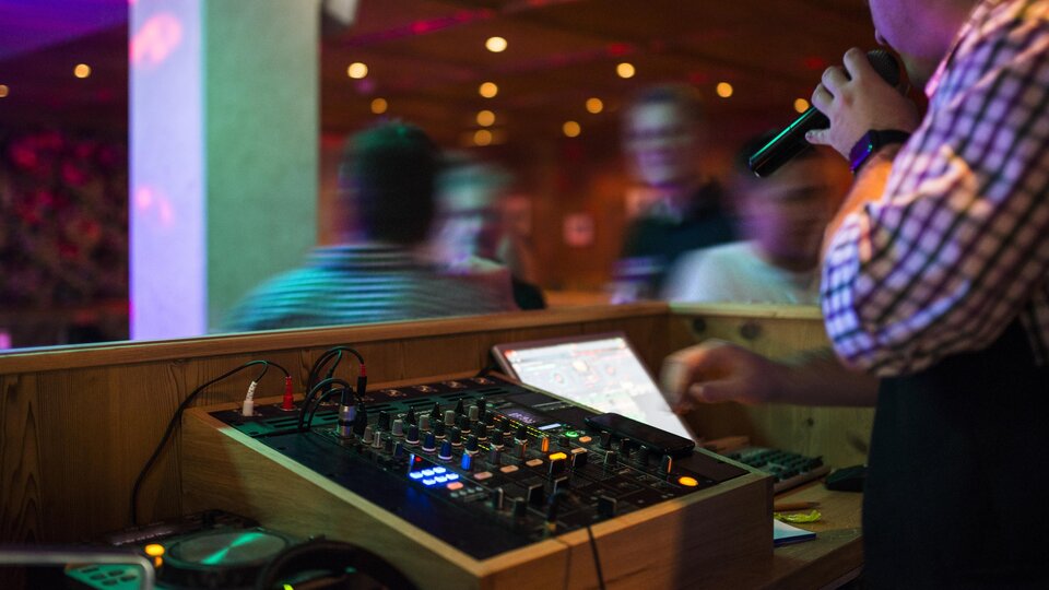 A DJ stands behind a mixing console and holds a microphone in hand. The colorful lights and the dancing crowd create a vibrant party atmosphere.