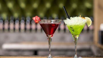 Two colorful cocktails are on a table. The left cocktail is red and the right one is green with a lime slice.