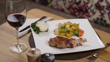 A juicy piece of meat on a white plate, accompanied by vegetables and mashed potatoes. Next to it, a glass of red wine on a wooden tray.