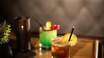 Two colorful cocktails on a table, one with an orange garnish and the other with a green color. Next to it lies a red drink menu and a candle creates a cozy atmosphere.