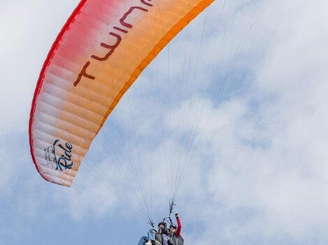 A paraglider hovers high in the sky. The canopy is orange and red, and the clouds are barely visible.