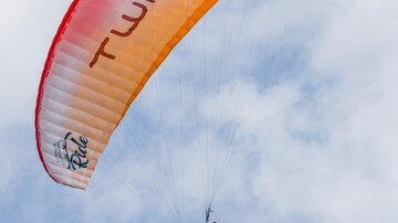 A paraglider hovers high in the sky. The canopy is orange and red, and the clouds are barely visible.