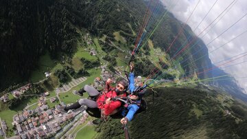 Three people paragliding over a green landscape. In the background, mountains and a village can be seen.