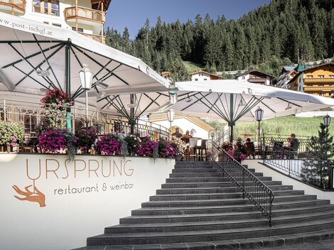 A cozy restaurant with terraces and blooming flowers. In the background, green mountains and traditional huts can be seen.