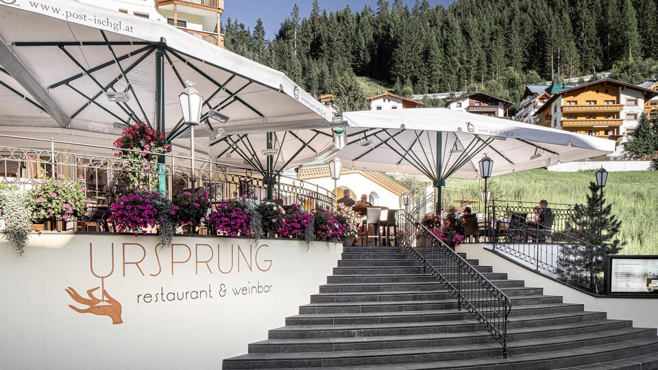 A cozy restaurant with terraces and blooming flowers. In the background, green mountains and traditional huts can be seen.