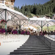 Ein gemütliches Restaurant mit Terrassen und blühenden Blumen. Im Hintergrund sind grüne Berge und traditionelle Hütten zu sehen.