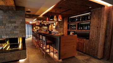 A modern bar with wood paneling and stylish light accents. The bar accommodates several guests and is well-stocked with drinks.