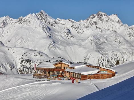 A cozy mountain cabin is situated in a snowy landscape. In the background, majestic mountains rise up under a clear sky.